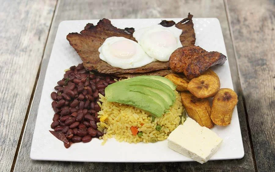 Plate with yellow rice and avocado on top accompanied by beans, meat, chorizo and plantain 